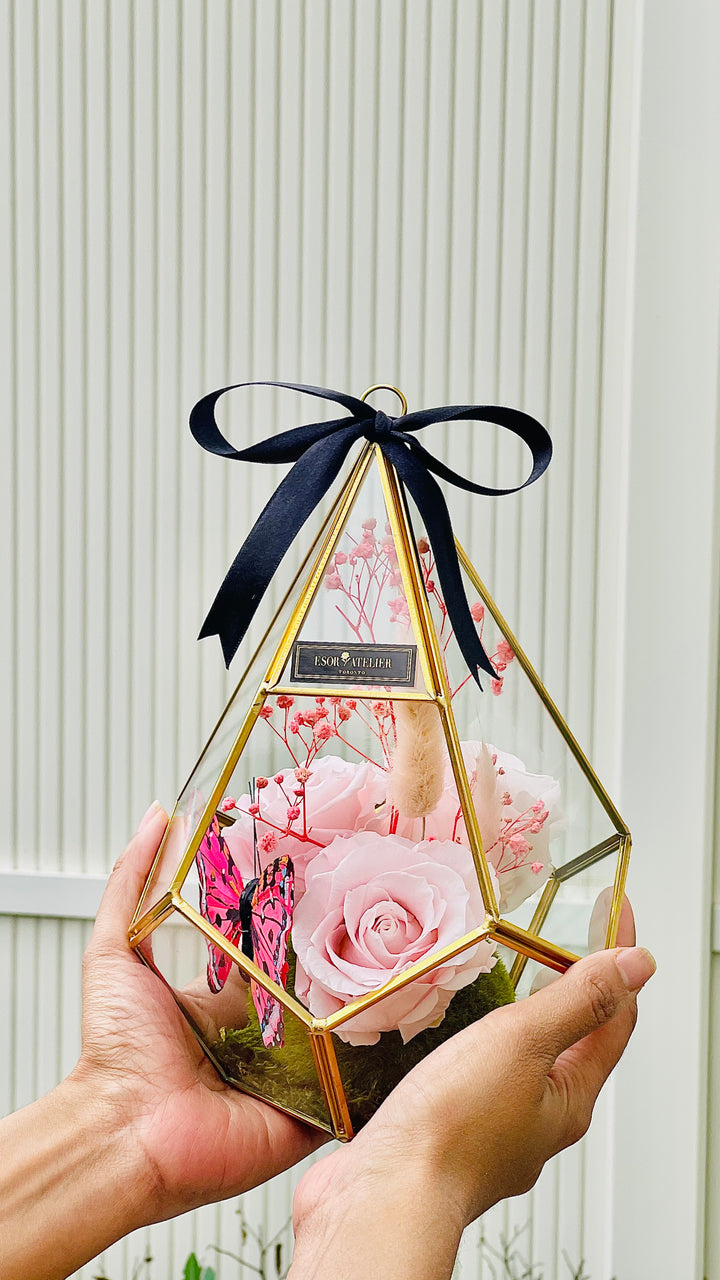 A geometric glass terrarium with preserved pink roses, green moss, and a black satin ribbon, held in two hands.