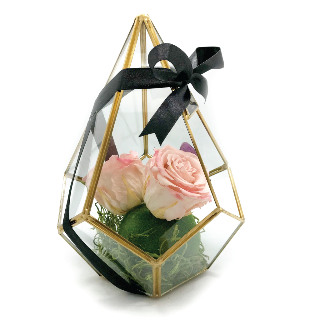 A geometric glass terrarium with preserved pink roses, green moss, and a black satin ribbon.